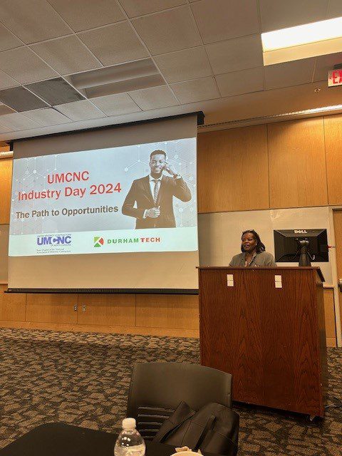 A person presenting at UMNC Industry Day 2024 event with the theme "The Path to Opportunities in General Contracting" displayed on the projector screen in the background.